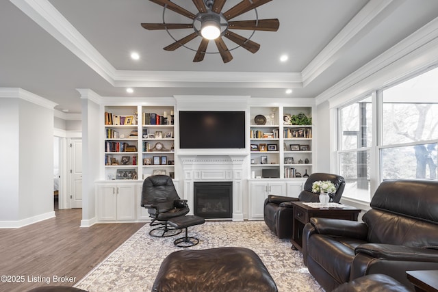 living area with a glass covered fireplace, ceiling fan, crown molding, wood finished floors, and a raised ceiling