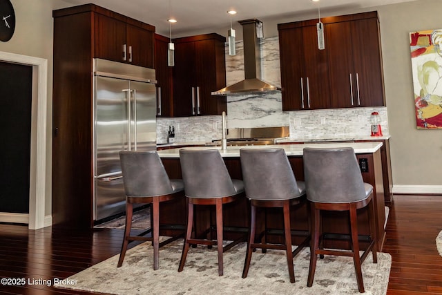 kitchen with wall chimney exhaust hood, dark wood finished floors, built in refrigerator, baseboards, and light countertops