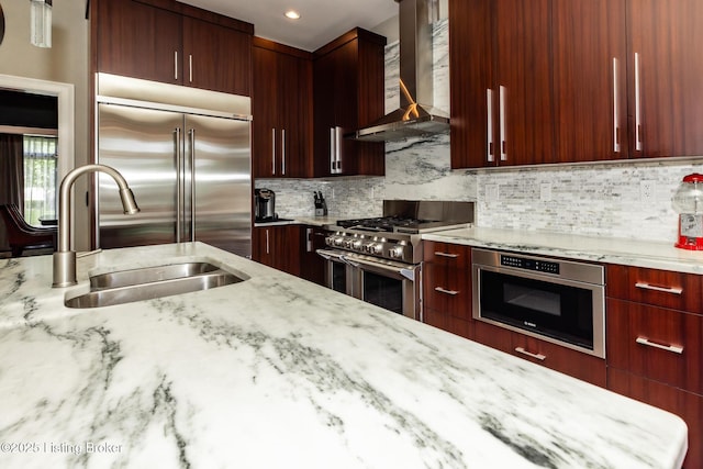 kitchen featuring a sink, high quality appliances, wall chimney exhaust hood, reddish brown cabinets, and light stone countertops