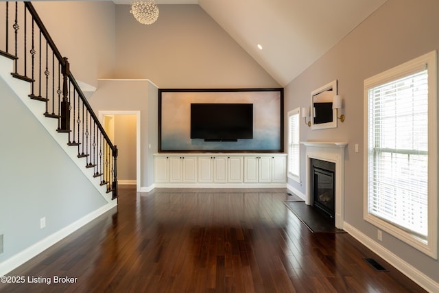 unfurnished living room featuring high vaulted ceiling, stairs, dark wood-style flooring, a glass covered fireplace, and baseboards