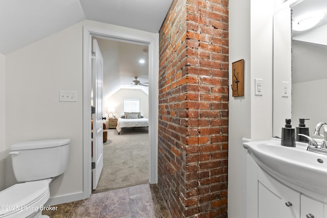 ensuite bathroom featuring a ceiling fan, toilet, brick wall, vanity, and vaulted ceiling
