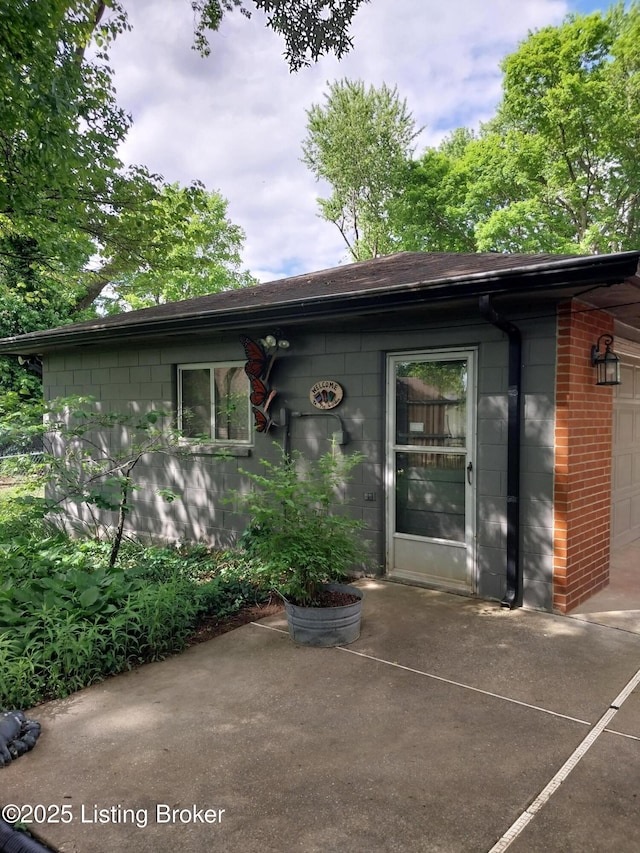 exterior space featuring a garage, concrete block siding, a shingled roof, a patio area, and brick siding