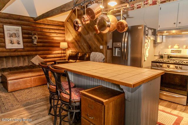 kitchen featuring tile countertops, under cabinet range hood, log walls, wood finished floors, and appliances with stainless steel finishes