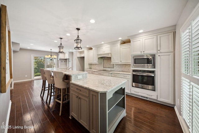 kitchen featuring appliances with stainless steel finishes, a center island with sink, decorative backsplash, open shelves, and a chandelier