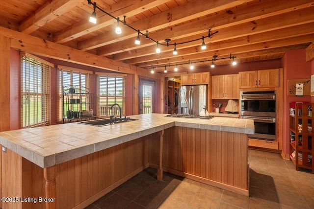 kitchen with appliances with stainless steel finishes, a sink, rail lighting, tile countertops, and wooden ceiling