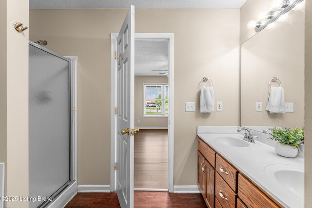 bathroom featuring baseboards, wood finished floors, a shower stall, double vanity, and a ceiling fan