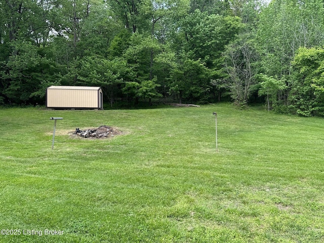 view of green lawn featuring an outdoor fire pit, a storage unit, an outdoor structure, and a wooded view