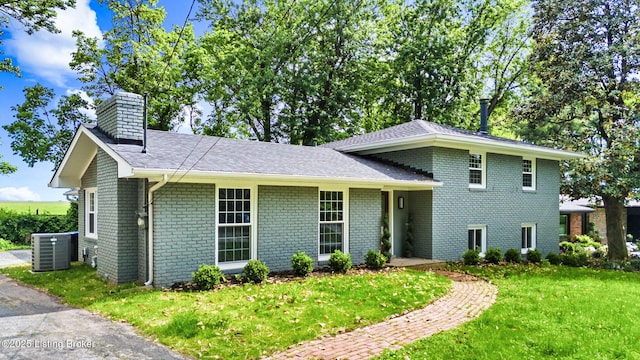 split level home with central air condition unit, brick siding, a chimney, a front yard, and roof with shingles
