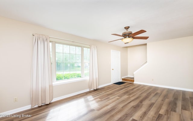 empty room with wood finished floors, baseboards, a ceiling fan, and stairway
