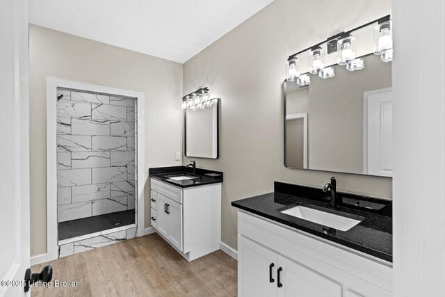 bathroom with a tile shower, two vanities, wood finished floors, and baseboards