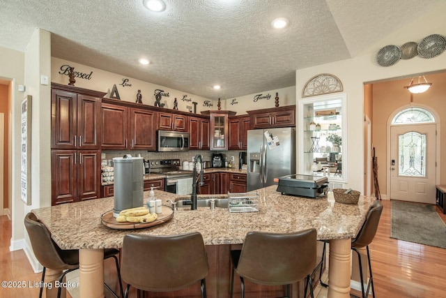 kitchen featuring appliances with stainless steel finishes, a textured ceiling, light wood finished floors, a breakfast bar, and glass insert cabinets