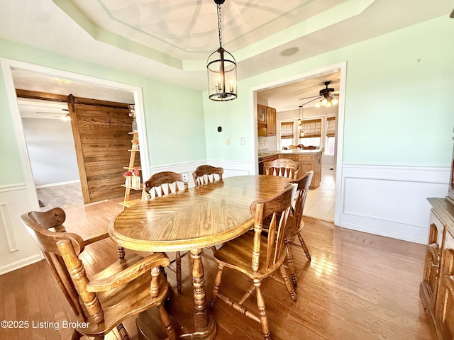 dining space featuring a tray ceiling, a wainscoted wall, light wood finished floors, a barn door, and a ceiling fan