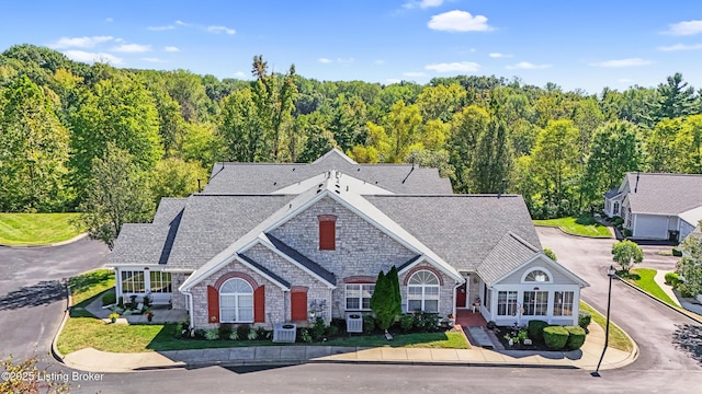view of front facade with stone siding, a wooded view, central AC unit, and roof with shingles