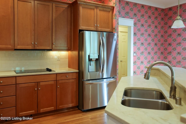 kitchen featuring stainless steel refrigerator with ice dispenser, wallpapered walls, a sink, black electric cooktop, and brown cabinets