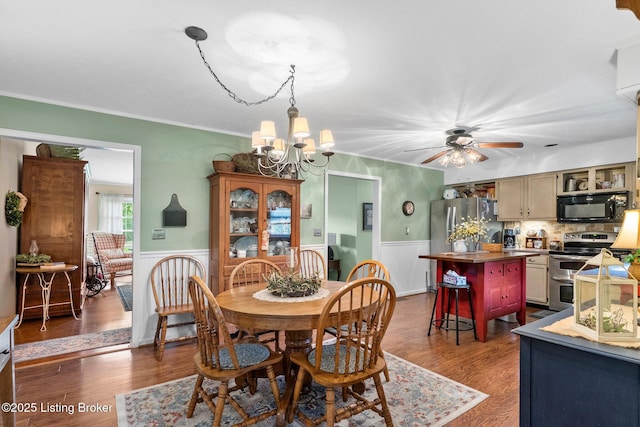 dining space with a wainscoted wall, a chandelier, ceiling fan, dark wood finished floors, and crown molding