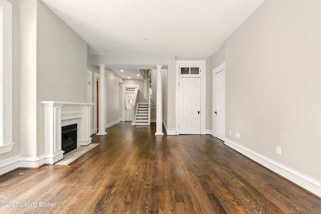 unfurnished living room featuring dark wood-style flooring, a fireplace with flush hearth, ornate columns, stairs, and baseboards