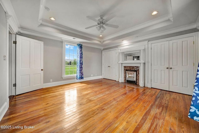 unfurnished living room featuring light wood-style flooring, a raised ceiling, a ceiling fan, a fireplace with flush hearth, and crown molding