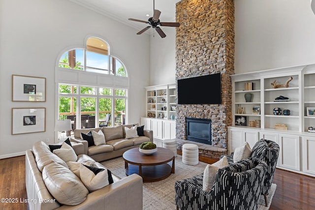 living room featuring a stone fireplace, a ceiling fan, dark wood-style flooring, a towering ceiling, and crown molding