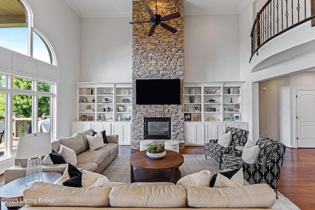 living area featuring ceiling fan, a stone fireplace, wood finished floors, a high ceiling, and crown molding