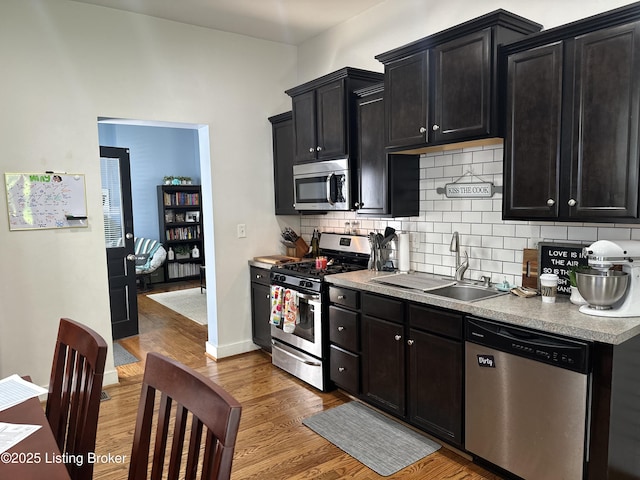 kitchen featuring appliances with stainless steel finishes, a sink, dark cabinets, wood finished floors, and decorative backsplash