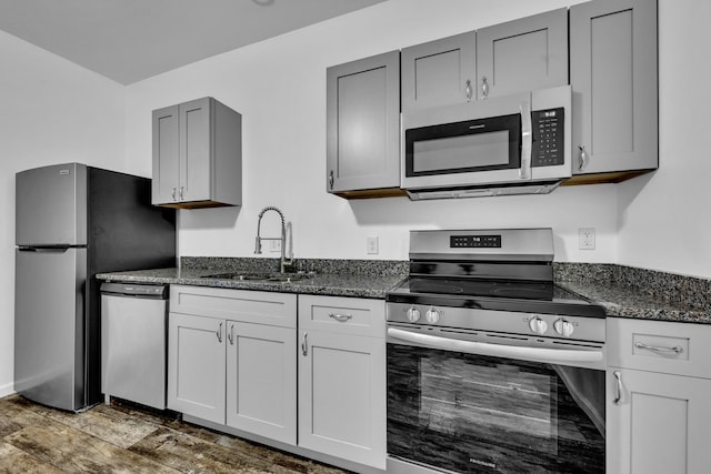 kitchen with stainless steel appliances, wood finished floors, a sink, gray cabinets, and dark stone counters