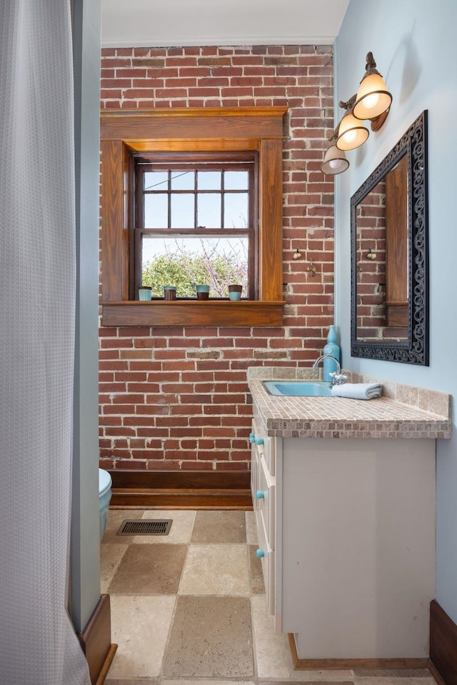 bathroom with stone tile flooring, visible vents, toilet, vanity, and brick wall