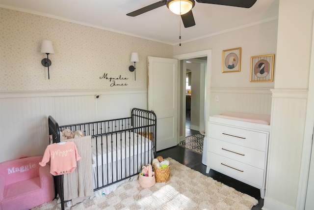 - bedroom featuring a nursery area, ornamental molding, wainscoting, dark wood-style flooring, and ceiling fan
