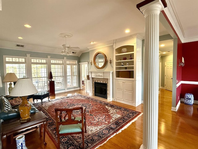 living room featuring crown molding, built in features, ceiling fan, wood-type flooring, and ornate columns