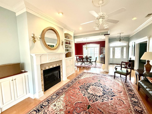 living room with ceiling fan with notable chandelier, a tile fireplace, ornamental molding, built in shelves, and ornate columns