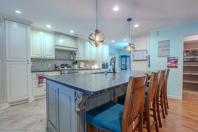 kitchen featuring electric range, sink, premium range hood, an island with sink, and decorative light fixtures