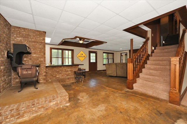  - unfurnished living room featuring a wood stove, concrete flooring, a ceiling fan, stairway, and a paneled ceiling