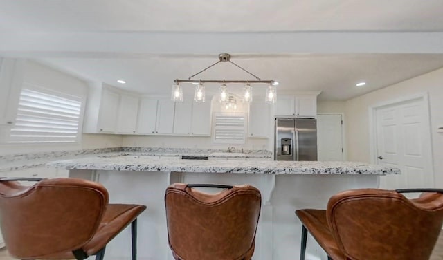 kitchen featuring white cabinetry, hanging light fixtures, stainless steel refrigerator with ice dispenser, light stone countertops, and a kitchen bar