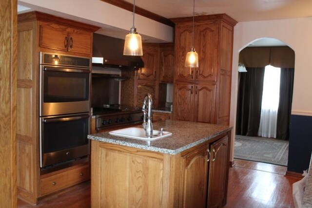 kitchen featuring sink, stainless steel double oven, dark wood-type flooring, pendant lighting, and a center island with sink