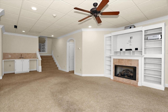 unfurnished living room with ceiling fan, a tiled fireplace, ornamental molding, light carpet, and built in shelves