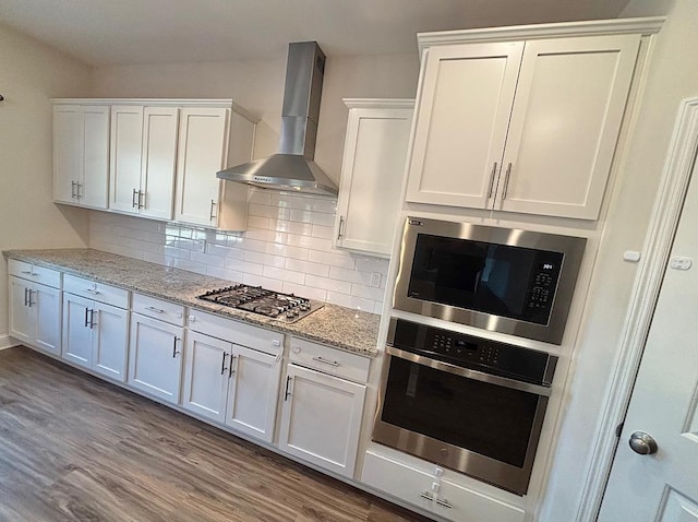 kitchen with light stone counters, stainless steel appliances, wall chimney range hood, hardwood / wood-style floors, and white cabinetry