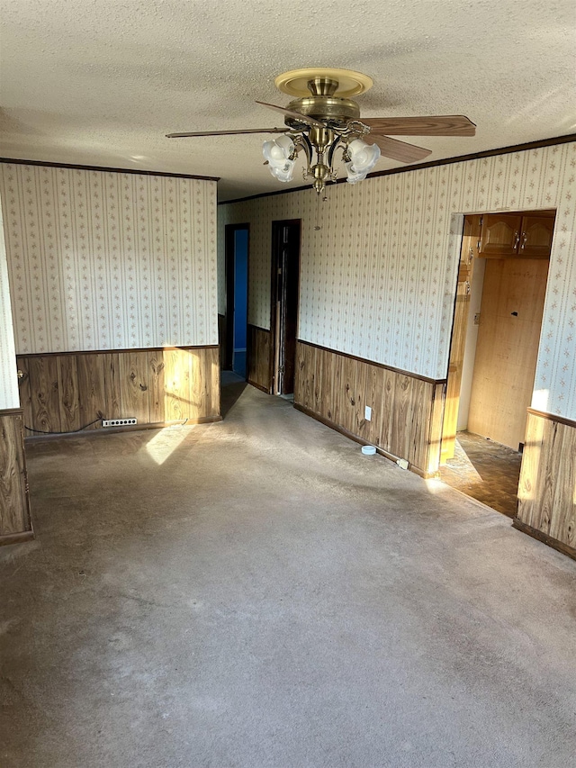 unfurnished room featuring wallpapered walls, wainscoting, ceiling fan, a textured ceiling, and crown molding