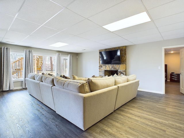 living area featuring a drop ceiling, a stone fireplace, baseboards, and wood finished floors