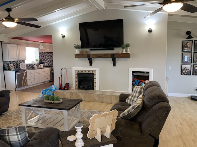 living area with vaulted ceiling with beams, ceiling fan, a tiled fireplace, and light wood-style flooring