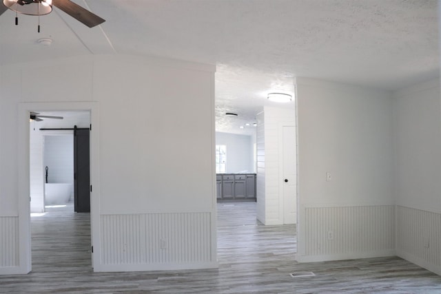 - empty room with light wood-style flooring, a barn door, a textured ceiling, wainscoting, and ceiling fan