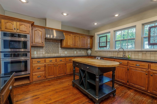  - kitchen with wall chimney exhaust hood, a sink, double oven, brown cabinetry, and recessed lighting