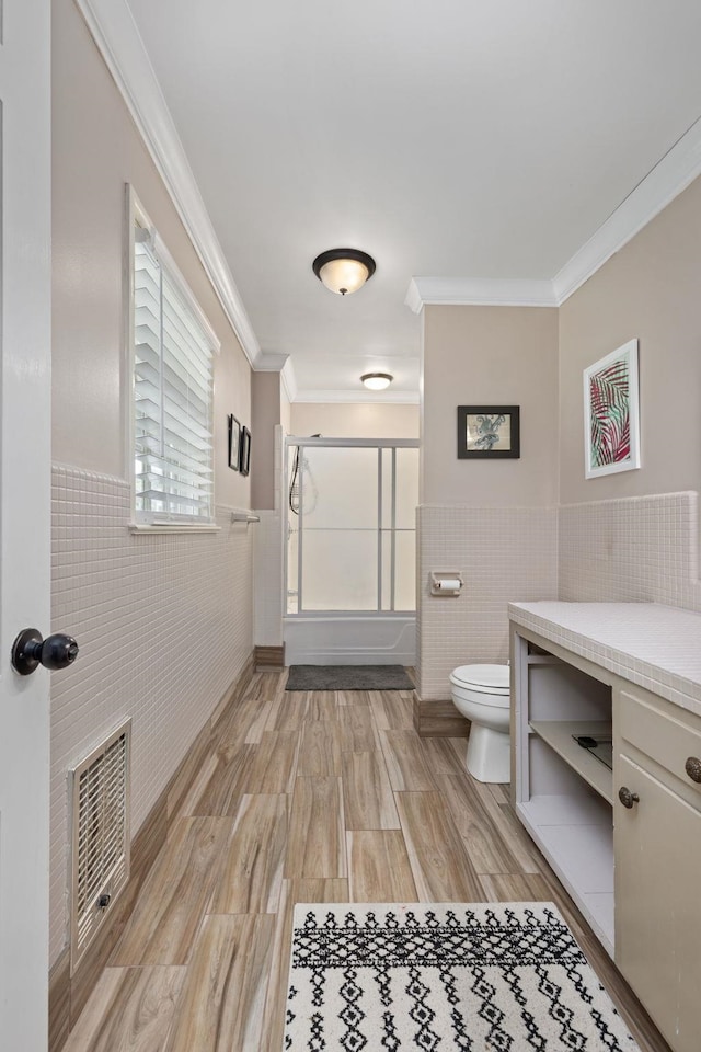  - bathroom featuring tile walls, wainscoting, toilet, ornamental molding, and bath / shower combo with glass door
