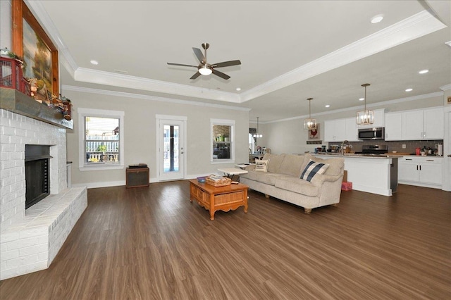 living room featuring crown molding, dark wood-type flooring, a fireplace, ceiling fan with notable chandelier, and a raised ceiling