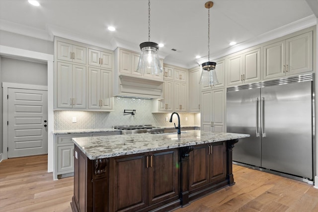 kitchen with decorative light fixtures, light wood-type flooring, built in fridge, light stone countertops, and a kitchen island with sink