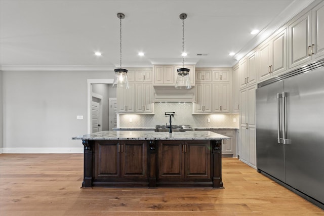 kitchen with light stone counters, stainless steel built in fridge, hanging light fixtures, and a center island with sink