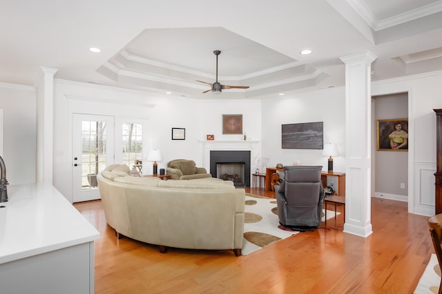 living room featuring ornamental molding, a tray ceiling, light wood-style floors, a fireplace, and ornate columns