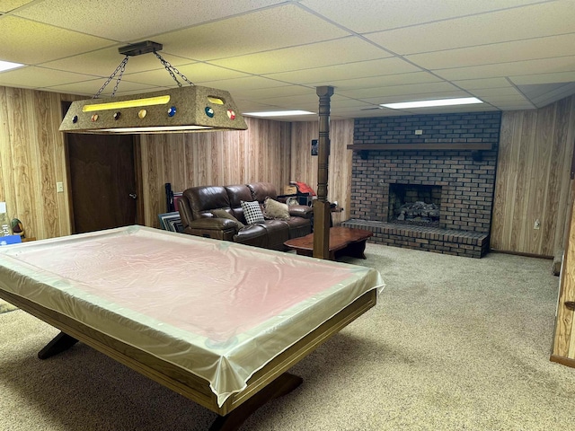 recreation room featuring carpet floors, wood walls, a drop ceiling, and a brick fireplace