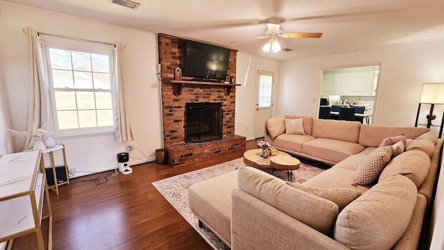 living room with a textured ceiling, visible vents, a ceiling fan, a brick fireplace, and dark wood-style floors