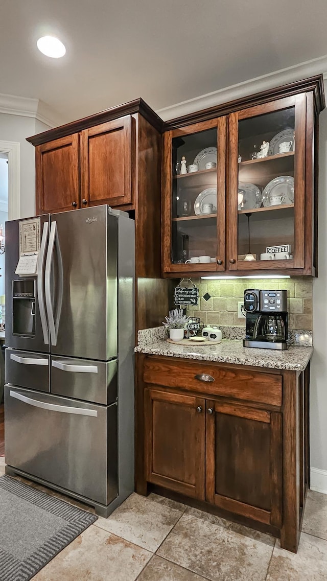 kitchen with stainless steel fridge, ornamental molding, backsplash, light stone counters, and glass insert cabinets
