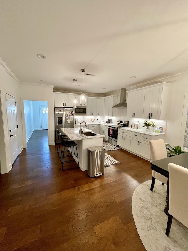 kitchen with appliances with stainless steel finishes, wall chimney range hood, a center island with sink, decorative light fixtures, and a breakfast bar area