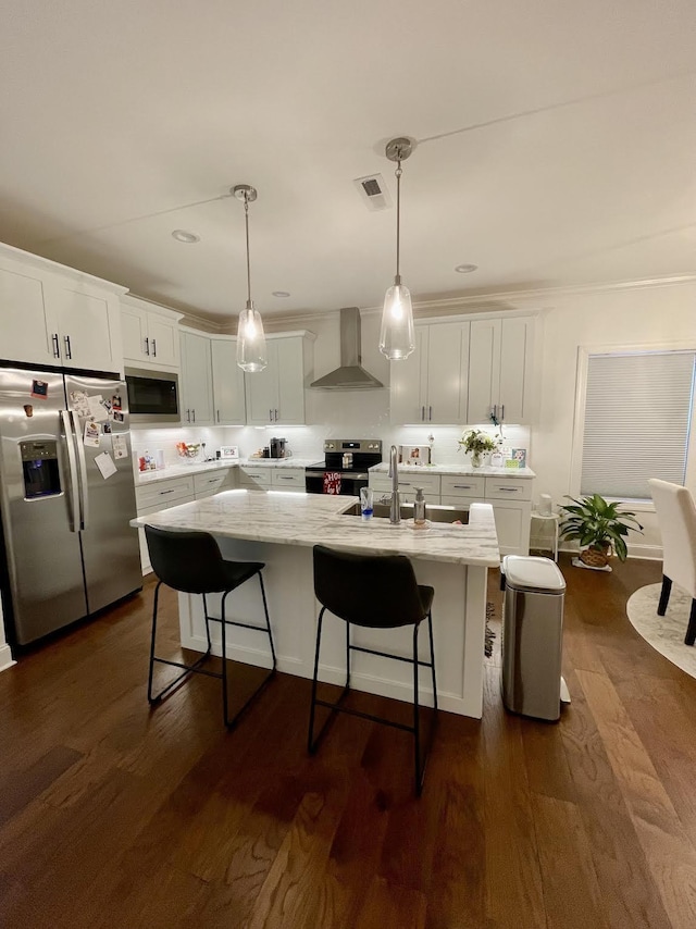 kitchen with wall chimney range hood, decorative light fixtures, a center island with sink, white cabinets, and appliances with stainless steel finishes